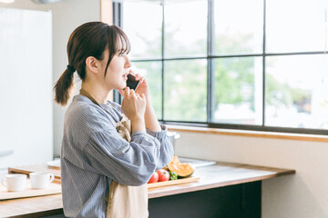 家のキッチン・ダイニングでスマホで電話しながら驚くアジア人女性
