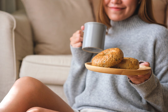 Closeup image of a beautiful young asian woman holding a plate of bagel while drinking hot coffee at home