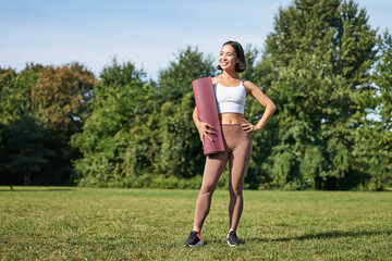 Portrait of young asian woman with rubber mat, standing on lawn and looking confident. Fitness...