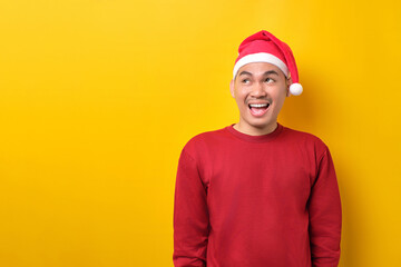 Happy young Asian man in Santa hat looking upwards with broad smile on yellow studio background. celebration Christmas holiday and New Year concept