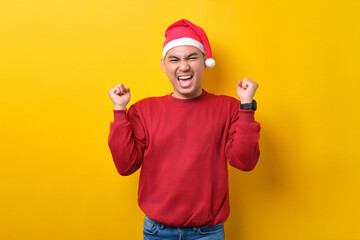 Naklejka premium Excited young Asian man in Santa hat raising hands up, celebrating success on yellow studio background. celebration Christmas holiday and New Year concept