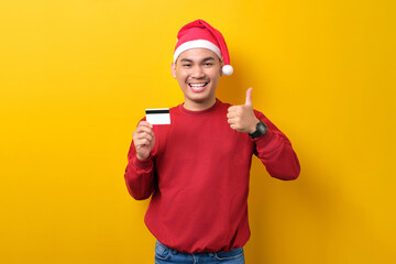 Cheerful young Asian man in Santa hat holding credit card and showing thumbs up on yellow studio background. celebration Christmas holiday and New Year concept