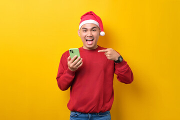 Excited young Asian man in Santa hat pointing finger at smartphone, recommending mobile application on yellow studio background. celebration Christmas holiday and New Year concept