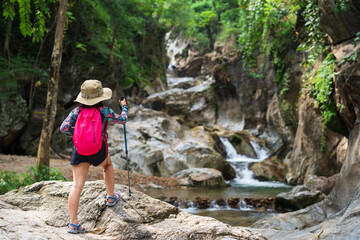 Obraz premium back of asian child or kid girl carry backpack and hiking by walking pole or trekking camping to standing on rocks and nature waterfall in forest for holiday adventure travel at lan sang national park