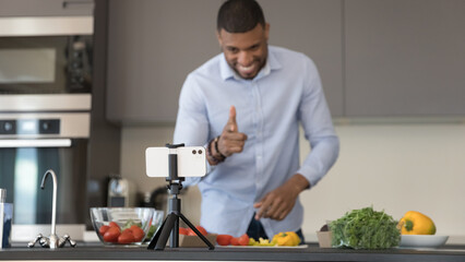 Cheerful African cook blogger shooting video post for blog, using mobile phone fixed on tripod stand, cooking salad in kitchen, speaking at webcam, promoting organic food healthy diet. Banner shot