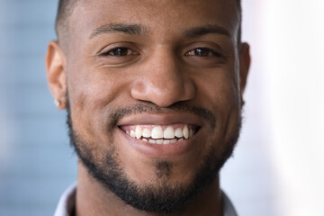Happy attractive young African guy face close up portrait. Handsome Black man looking at camera with toothy smile, showing healthy white perfect teeth, promoting dental service, male beauty care