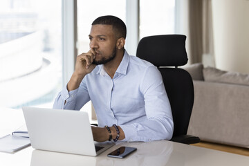 Naklejka premium Happy millennial African freelance businessman looking away from office workplace, sitting at laptop computer on work table, thinking over project future vision, task completion