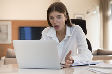 Shocked millennial freelance employee girl looking at laptop with open mouth surprised with bad...