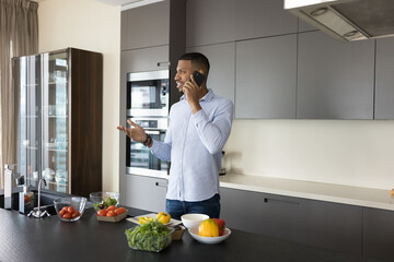 Positive handsome young African chef guy making call on mobile phone at kitchen table with organic ingredients, talking on cellphone, cooking salad, preparing healthy dinner, asking recipe