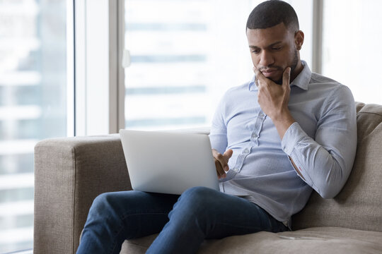 Focused thoughtful Black freelancer man using laptop on home sofa, watching online video, reading message, notice, thinking, feeling doubt, making decision, touching chin