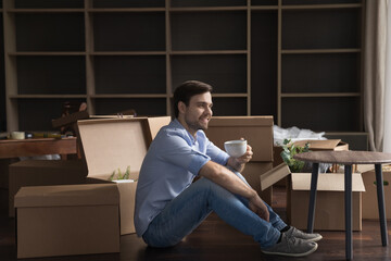 Happy carefree property buyer man enjoying moving into new home, sitting on heating floor among cardboard boxes, drinking tea, taking coffee break, holding hot mug, looking away, smiling