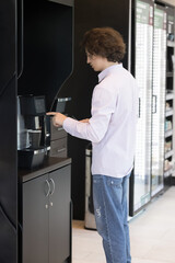 Naklejka na ściany i meble Vertical view, young man standing in modern office workspace during lunch break prepare morning hot coffee or tea beverage using professional vending machine indoor. Food and drink industry, lifestyle