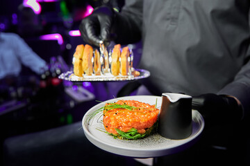 The waiter is holding Salmon tartare with avocado on a white plate in a restaurant. Close-up, selective focus