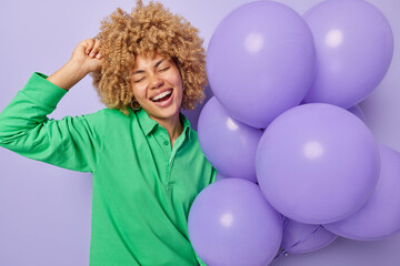 Festive occasion partying concept. Cheerful curly haired woman laughs gladfully holds bunch of inflated balloons enjoys birthday celebration smiles broadly keeps eyes closed isolated over purple wall