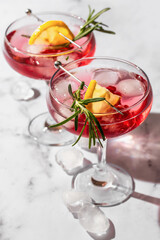 Two glasses of Pomegranate iced cocktail with rosemary and lemon on white marble background isolated with clear shadows in hard light close up. Xmas drink party concept