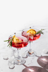 Two glasses of Pomegranate iced cocktail with rosemary and lemon on white marble background isolated with clear shadows in hard light with text space. Xmas drink party concept