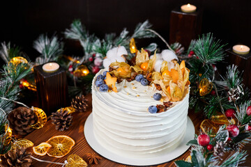 Christmas cake made of chocolate base with cream, decorated with blueberries and physalis against the backdrop of Christmas decor