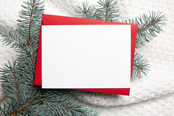 Christmas greeting card mockup with red envelope and fir tree branches on white knitted clothes background, top view, flat lay