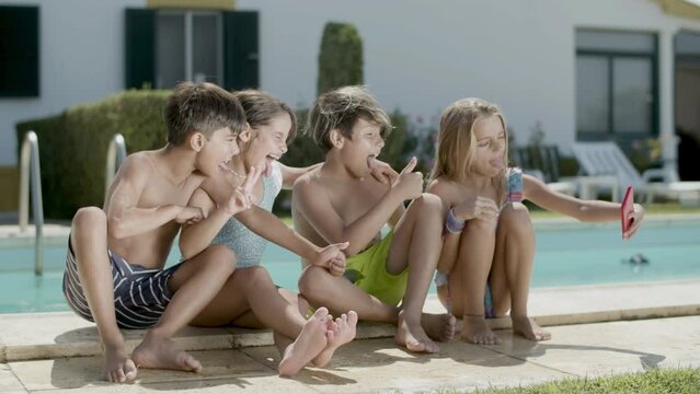 Children sitting on poolside taking selfie on sunny day. Cheerful boys and girls making selfie showing tongues to camera, holding thumbs up. Childhood, joy, modern technology concept.