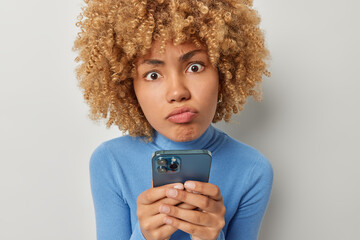 Portrait of upset dissatisfied woman purses lips looks sadly at camera feels worried about something holds cellphone in hands dressed in casual blue turtleneck isolated over grey background.