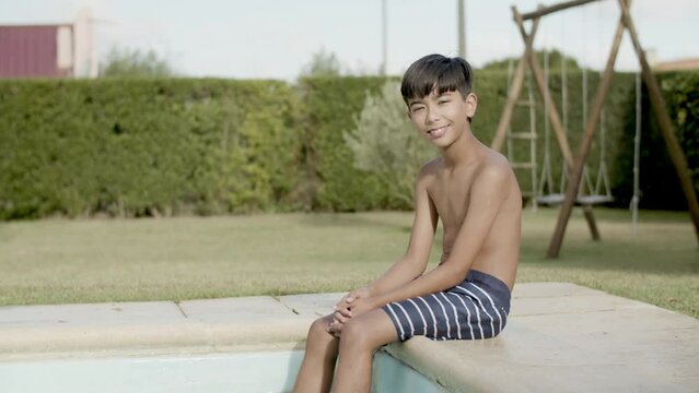 Asian boy sitting on pool edge smiling, dangling feet in water. Boy in swimming trunks enjoying his vacation time outside. Playground with swings in background. Summer, childhood concept. 