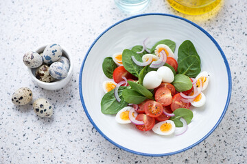 Plate with spinach, cherry tomatoes and quail eggs salad, elevated view on a light-grey granite background, horizontal shot