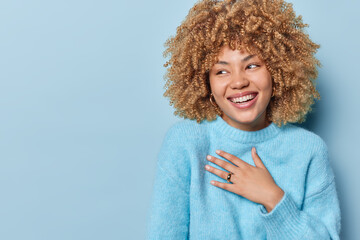 Horizontal shot of happy sincere woman with curly hair keeps hand on chest smiles broadly expresses...