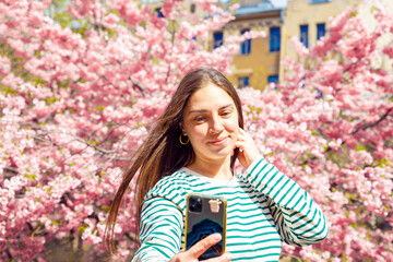 Making selfie woman on smartphone on cherry blossom sakura background. Young smiling european white female is taking selfie portrait with mobile phone. Summer Spring headshot lifestyle portrait