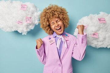 Overjoyed female entrepreneur with curly blonde hair dressed formally clenches fists exclaims...
