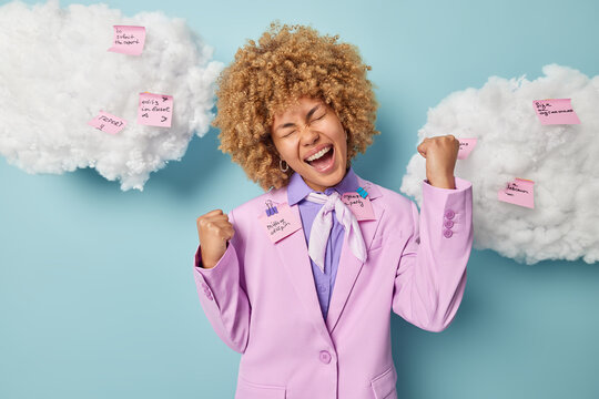 Overjoyed female entrepreneur with curly blonde hair dressed formally clenches fists exclaims loudly feels like winner isolated on blue wall white clouds overhead sticky notes written tasks to do