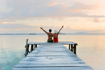 couple of tourists with arms in the sky sitting on the wooden jetty watching the sunset - travel in...