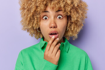 Cropped shot of amazed curly haired young woman stares with widely opened eyes and mouth reacts to something shocking wears green jumper isolated over purple background. Human reactions concept