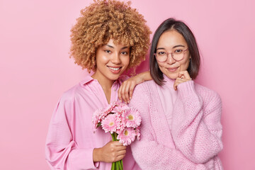 Two pleased women of different race stand next to each other in casual clothes hold beautiful bouquet of gerbera flowers smile pleasantly enjoy holiday time isolated over pink studio background.