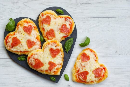 Heart shaped mini pizzas with heart shaped pepperoni on heart shaped slate plate with white wood background.Creative art food idea for celebrate Valentine or Mother day.Top view.Copy space