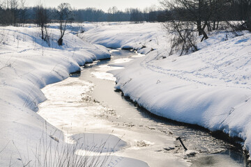 Winter landscape. Small river with ice and snow-covered field on a bright sunny day