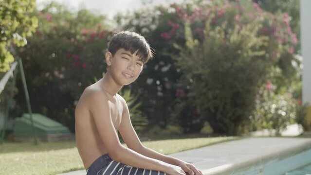 Happy boy sitting on poolside, smiling and squinting from sun. Slender Asian boy having fun by the pool, splashing water with his feet. Side view, medium shot. Childhood, vacation concept.