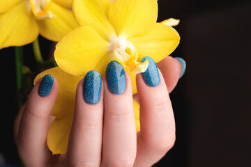 Female hand with beautiful manicure - blue glitter nails with yellow phalaenopsis orchid flower. Nail care concept