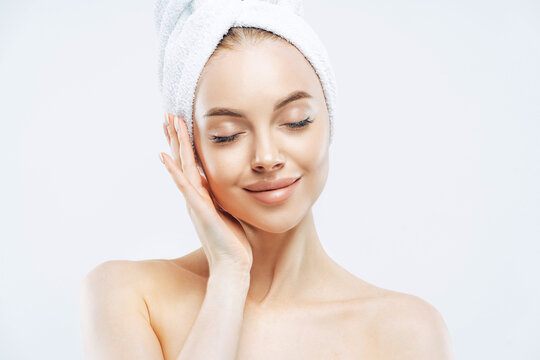 Pretty calm woman with wrap towel on head, closes eyes, enjoys softness of skin, undergoes skin care treatment, stands bare shoulders indoor, relaxed after bath, isolated over white background