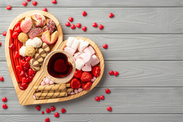 Valentine's Day concept. Top view photo of wooden heart shaped serving tray with sweets glass of drinking candies and cookies on grey wooden table background with copyspace