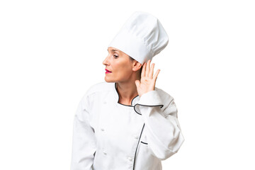 Middle-aged chef woman over isolated background listening to something by putting hand on the ear