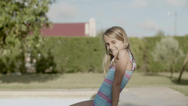 Young girl sitting on poolside, smiling and squinting from sun. Cute Caucasian girl spending vacation time by pool, dangling feet in water. Medium shot, side view. Childhood, summer concept.