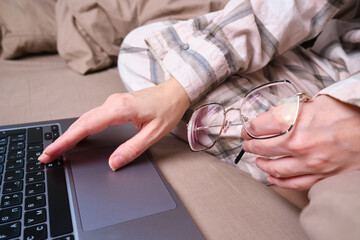 Young woman working on a laptop in glasses in bed. Glasses for vision in hands close-up. Freelance work from home