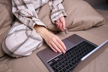 Young woman working on a laptop in glasses in bed. Glasses for vision in hands close-up. Freelance work from home