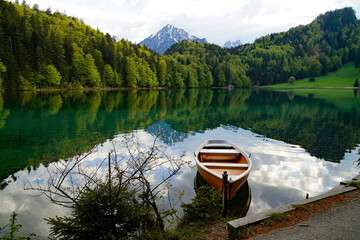 a boat resting on the emerald-green waters of lake Alatsee in Fuessen with the snowy Bavarian Alps reflected in the calm water and the lush green spring forest in the background, Bavaria, Germany