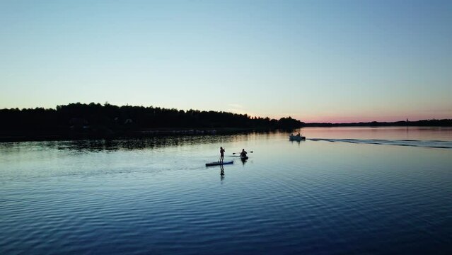 Woman and two kids enjoy on a supboard. Outdoor activity on a lake during midsummer blue hour sunset warm colors in sweden. People in a motor boat passes the sup surfers, arc shot low angle view