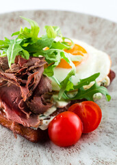 Fried eggs with arugula and thin slices of beef on rye bread toast and cherry tomatoes.
