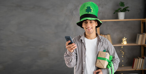 smiling young man in leprechaun hat holding gift box, using mobile phone, St.Patrick's day promotion sale app on smartphone