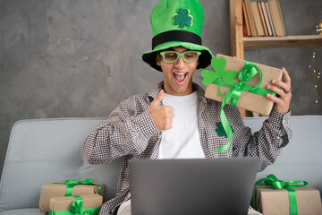 Young happy man having video call over laptop while celebrating St.Patrick's Day alone at home. Thumbs up gesture