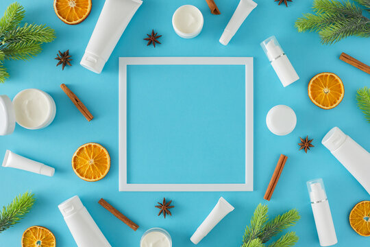 Natural season cosmetics concept. Flat lay photo of cosmetic bottles without label, dried orange slices, cinnamon sticks and fir branches on pastel blue background with frame.