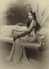 Portrait flapper woman in twenties old style sitting on sofa, couch. Long evening retro dress...